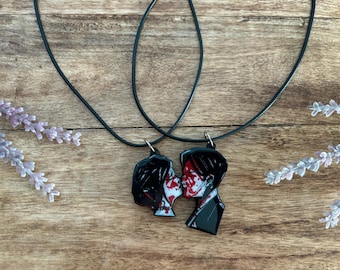 May include: Two black pendant necklaces featuring a couple kissing with red accents. The pendants hang from black cords. The image is set against a wooden background with lavender sprigs.