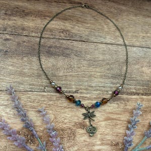 Puede incluir: Un delicado collar con una cadena de bronce y cuentas coloridas en tonos morado, azul y &aacute;mbar. Un dije de lib&eacute;lula y detalles de botones cuelgan del centro. El collar descansa sobre una superficie de madera con ramitas de lavanda.