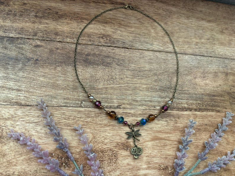 Puede incluir: Un delicado collar con una cadena de bronce y cuentas coloridas en tonos morado, azul y &aacute;mbar. Un dije de lib&eacute;lula y detalles de botones cuelgan del centro. El collar descansa sobre una superficie de madera con ramitas de lavanda.