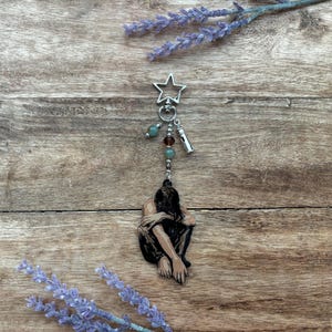 Puede incluir: Un llavero plateado con una parte superior en forma de estrella, cuentas verdes y marrones y un pequeño dije de linterna plateada. El llavero presenta una figura de una persona en posición sentada. El fondo es una superficie de madera con ramitas de lavanda.