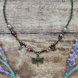 May include: A bronze necklace with a dragonfly pendant, featuring small pink flowers and star charms. The necklace is made with brown and blue beads.