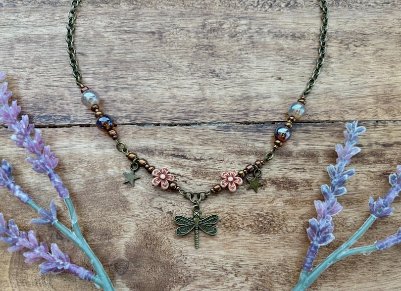 May include: A bronze necklace with a dragonfly pendant, featuring small pink flowers and star charms. The necklace is made with brown and blue beads.
