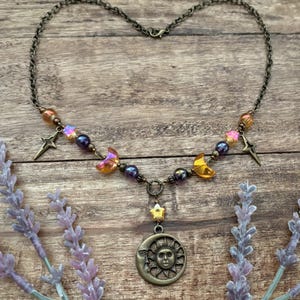 Op de afbeelding: Een hemelsblauwe ketting met een bronzen hanger. De hanger heeft een zon- en maanontwerp, aangevuld met ster- en halve maan bedels. De ketting bevat kleurrijke kralen en is geplaatst tegen een houten achtergrond.