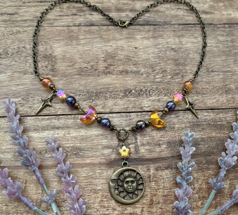Op de afbeelding: Een hemelsblauwe ketting met een bronzen hanger. De hanger heeft een zon- en maanontwerp, aangevuld met ster- en halve maan bedels. De ketting bevat kleurrijke kralen en is geplaatst tegen een houten achtergrond.