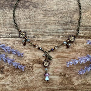 May include: A bronze necklace with floral and bead details, and a mushroom pendant. The necklace is set against a wooden background with lavender sprigs. The chain is delicate.