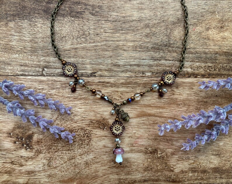 May include: A bronze necklace with floral and bead details, and a mushroom pendant. The necklace is set against a wooden background with lavender sprigs. The chain is delicate.