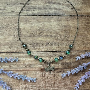 May include: A bronze necklace featuring iridescent green and blue beads and a dragonfly charm. The necklace is set against a wooden background, with sprigs of lavender.