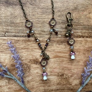 May include: A bronze-coloured necklace and keychain set with floral and mushroom charms. The necklace has a chain with flower-shaped beads and a mushroom pendant. The keychain has a lobster clasp and a mushroom charm. The set is on a wooden surface.