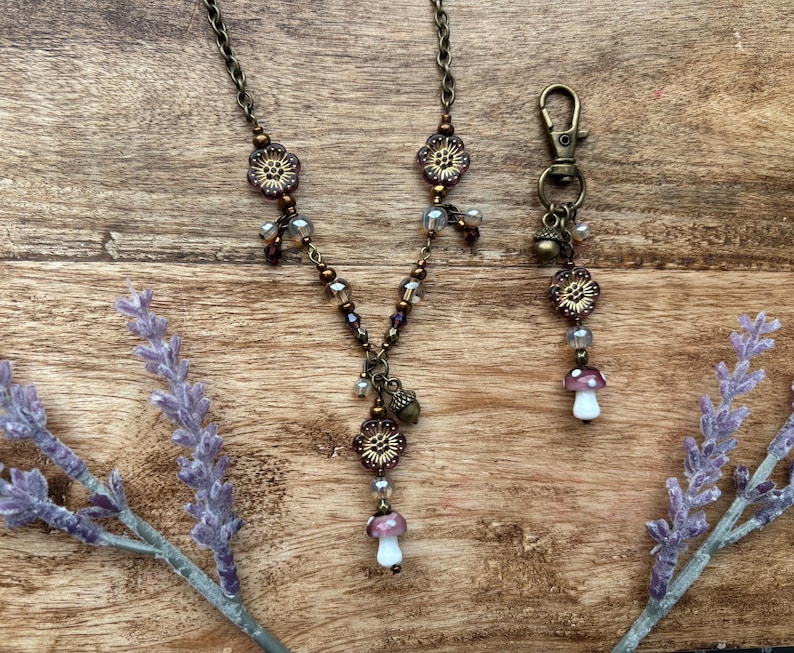 May include: A bronze-coloured necklace and keychain set with floral and mushroom charms. The necklace has a chain with flower-shaped beads and a mushroom pendant. The keychain has a lobster clasp and a mushroom charm. The set is on a wooden surface.