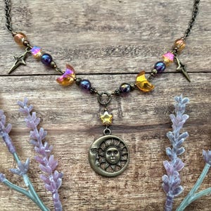 Op de afbeelding: Een ketting met een hemels thema met een bronzen zon- en maan hanger. De ketting bevat ster- en halve maan bedels, iriserende en paarse kralen. De ketting wordt gepresenteerd op een houten oppervlak met lavendel takjes.
