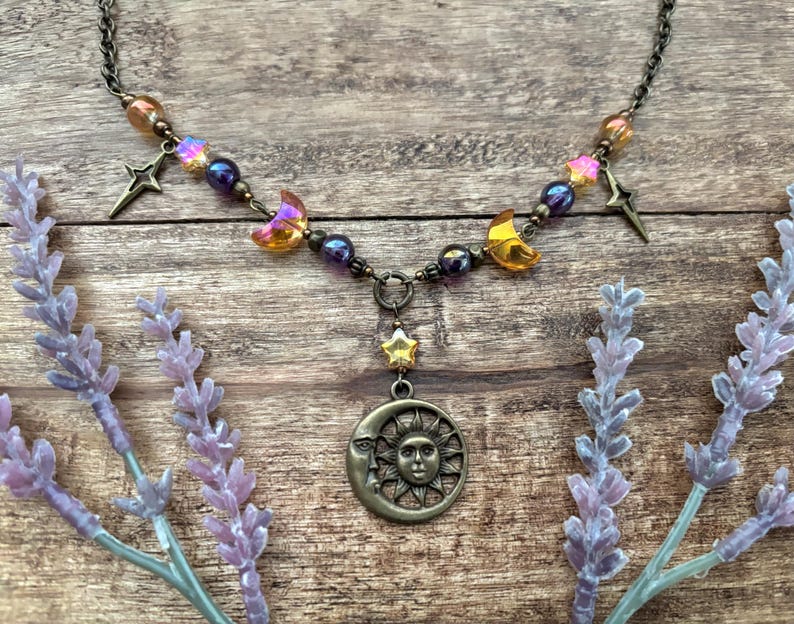 Op de afbeelding: Een ketting met een hemels thema met een bronzen zon- en maan hanger. De ketting bevat ster- en halve maan bedels, iriserende en paarse kralen. De ketting wordt gepresenteerd op een houten oppervlak met lavendel takjes.