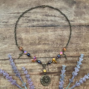 Op de afbeelding: Een ketting met een bronzen ketting en hanger met een zon-gezicht ontwerp. De ketting is versierd met iriserende kralen, sterbedels en halve maan bedels. Lavendel takken omlijsten de ketting.