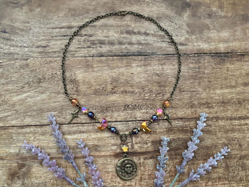 Op de afbeelding: Een ketting met een bronzen ketting en hanger met een zon-gezicht ontwerp. De ketting is versierd met iriserende kralen, sterbedels en halve maan bedels. Lavendel takken omlijsten de ketting.