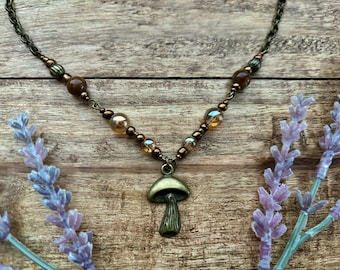 Puede incluir: Un collar de tono bronce con un colgante de seta y una cadena de cuentas marrones y ámbar. El collar está sobre un fondo de madera, con ramitas de lavanda a cada lado. Una joya de inspiración natural.