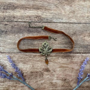 May include: A brown velvet choker necklace featuring a central antique brass-toned metal leaf charm. The leaf is accented with a small, dangling amber-coloured bead. The choker includes a metal lobster claw clasp and an adjustable chain extension.