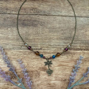 Puede incluir: Un delicado collar con un colgante de lib&eacute;lula. El collar presenta una cadena de bronce adornada con cuentas de colores en tonos morado, azul y &aacute;mbar. El colgante de lib&eacute;lula cuelga debajo de dos peque&ntilde;os amuletos de calavera. Ramas de lavanda enmarcan el collar sobre un fondo de madera.