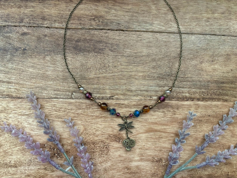 Puede incluir: Un delicado collar con un colgante de lib&eacute;lula. El collar presenta una cadena de bronce adornada con cuentas de colores en tonos morado, azul y &aacute;mbar. El colgante de lib&eacute;lula cuelga debajo de dos peque&ntilde;os amuletos de calavera. Ramas de lavanda enmarcan el collar sobre un fondo de madera.