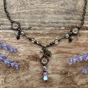 May include: A handmade necklace with a bronze chain and floral details. The necklace features a mushroom pendant with a white stem and a purple cap. Clear and purple beads are incorporated into the design.