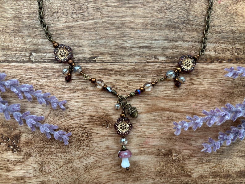 May include: A handmade necklace with a bronze chain and floral details. The necklace features a mushroom pendant with a white stem and a purple cap. Clear and purple beads are incorporated into the design.