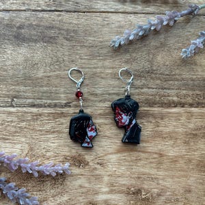 May include: A pair of black and red earrings. Each earring features a profile of a person with red accents on the face and a black outfit. The earrings hang from silver-colored hooks and have a red bead detail.