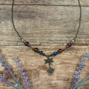 Puede incluir: Un delicado collar con un colgante de lib&eacute;lula y cuentas de colores. El collar presenta una cadena de bronce y un dije de lib&eacute;lula con tres peque&ntilde;os dijes de bot&oacute;n. Las cuentas son en tonos morado, azul y &aacute;mbar.