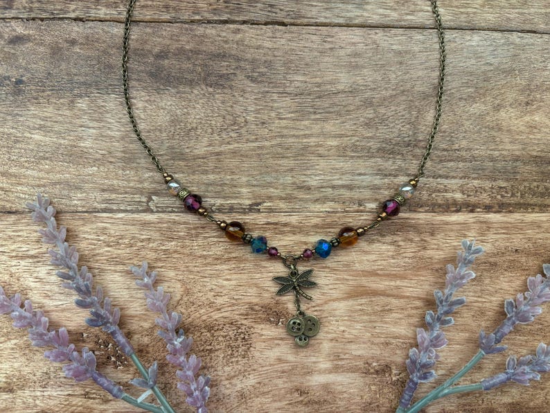 Puede incluir: Un delicado collar con un colgante de lib&eacute;lula y cuentas de colores. El collar presenta una cadena de bronce y un dije de lib&eacute;lula con tres peque&ntilde;os dijes de bot&oacute;n. Las cuentas son en tonos morado, azul y &aacute;mbar.