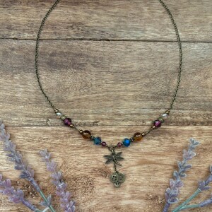 Puede incluir: Un delicado collar con un colgante de lib&eacute;lula y cuentas de colores. El collar presenta una cadena de color bronce y un dije de lib&eacute;lula con dos peque&ntilde;os dijes de bot&oacute;n. Las cuentas son en tonos morado, azul y &aacute;mbar.