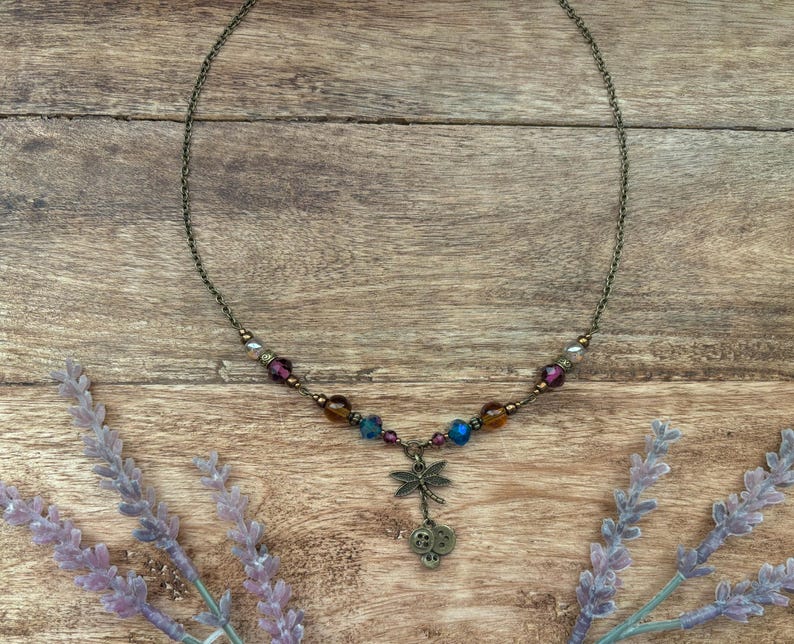 Puede incluir: Un delicado collar con un colgante de lib&eacute;lula y cuentas de colores. El collar presenta una cadena de color bronce y un dije de lib&eacute;lula con dos peque&ntilde;os dijes de bot&oacute;n. Las cuentas son en tonos morado, azul y &aacute;mbar.