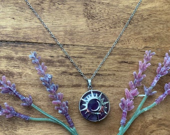 Op de afbeelding: Een zilveren ketting met een hanger met een zon- en maandesign. De hanger is gemaakt van metaal en heeft een paarse steen in het midden.