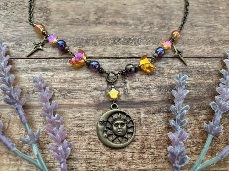 Op de afbeelding: Een ketting met een hemels thema met een bronzen hanger met een zon- en maanontwerp. De ketting heeft een ketting met ster- en halve maan bedels, en iriserende en paarse kralen. De ketting is geplaatst tegen een houten achtergrond met lavendel takjes.