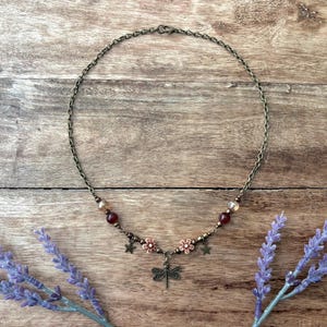 May include: A bronze-toned chain necklace featuring a central dragonfly charm, two small flower charms, two star charms, and dark red and translucent beads. The necklace is secured with a lobster claw clasp and has a delicate, symmetrical design.