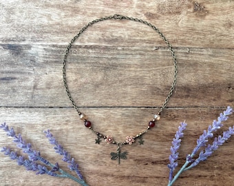 May include: A bronze-toned chain necklace featuring a central dragonfly charm, two small flower charms, two star charms, and dark red and translucent beads. The necklace is secured with a lobster claw clasp and has a delicate, symmetrical design.