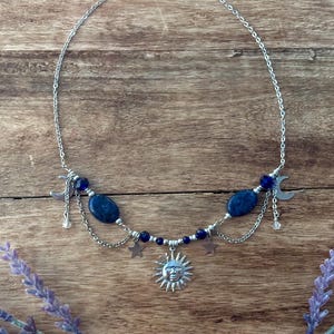 May include: A silver-toned necklace featuring a central sun pendant, two oval blue beads, small star charms, and crescent moon accents. The design includes delicate silver chains and dark blue faceted beads arranged symmetrically along the metal chain.