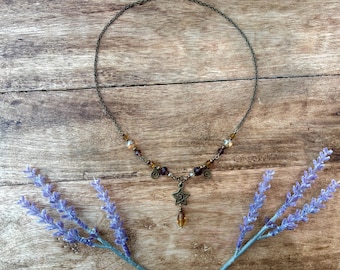 May include: A necklace featuring a thin dark metal chain, accented with small amber and dark brown beads, and a central bronze-toned star charm with a spiral design, finished with a single dangling amber-colored teardrop bead at the bottom.