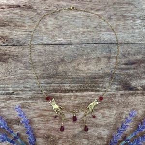Può includere: Una collana color oro con due ciondoli a forma di mano simmetrici, piccoli dettagli a stella e diverse perline pendenti rosso scuro a forma di goccia. La catenina è sottile e metallica, collegata agli elementi decorativi da un moschettone.