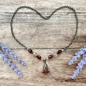 May include: A delicate necklace with a dark chain forming a heart shape. The necklace features burgundy and pink beads, and a flower pendant. Lavender sprigs frame the jewelry on a wooden surface.