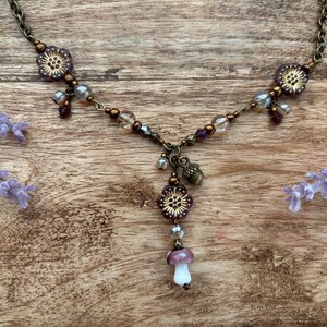 May include: A detailed necklace with a bronze chain and floral and mushroom pendants. The necklace features purple, clear, and bronze beads, with a small acorn charm. The mushroom pendant has a white stem and a purple cap. Lavender sprigs are in the background.