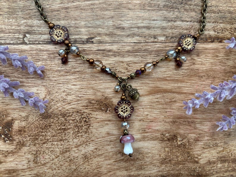 May include: A detailed necklace with a bronze chain and floral and mushroom pendants. The necklace features purple, clear, and bronze beads, with a small acorn charm. The mushroom pendant has a white stem and a purple cap. Lavender sprigs are in the background.