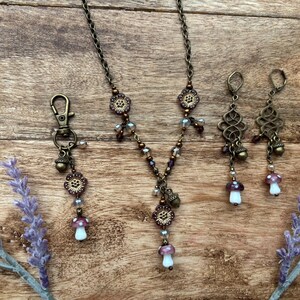 May include: An antique bronze-toned jewellery set comprising a necklace, earrings, and a keychain. The necklace and keychain feature floral and mushroom charms, while the earrings have an ornate design. The set is accented with purple and clear beads.