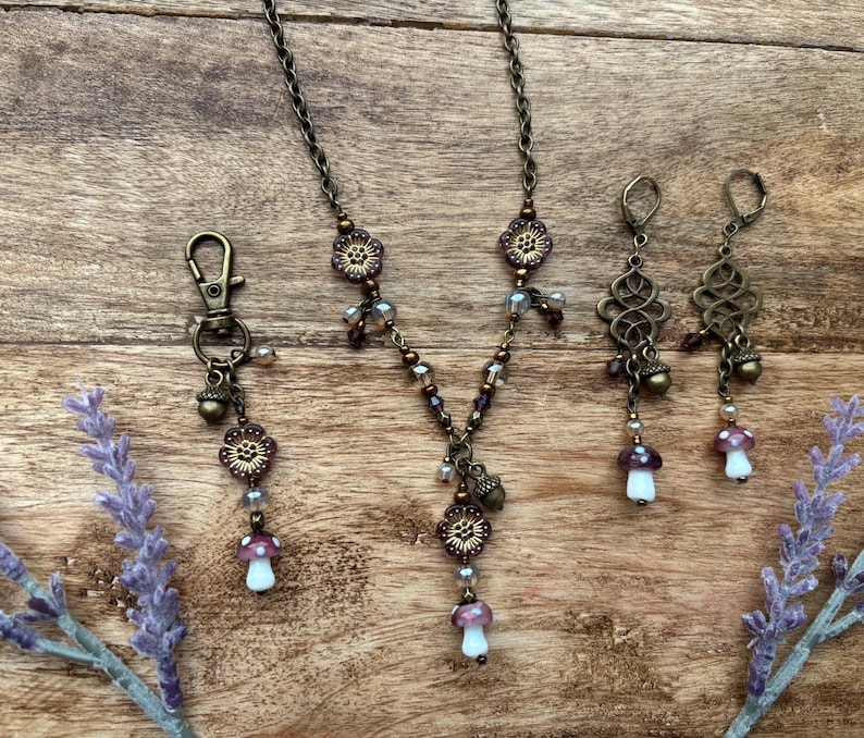 May include: An antique bronze-toned jewellery set comprising a necklace, earrings, and a keychain. The necklace and keychain feature floral and mushroom charms, while the earrings have an ornate design. The set is accented with purple and clear beads.