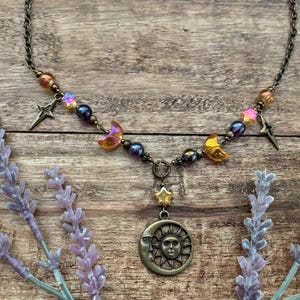 Op de afbeelding: Een ketting met hemels thema met een bronzen ketting en hanger. De hanger heeft een zon- en maanontwerp, aangevuld met ster- en maanbedels en iriserende kralen in oranje, paars en goud. De ketting wordt gepresenteerd op een houten oppervlak.