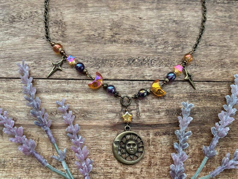 Op de afbeelding: Een ketting met hemels thema met een bronzen ketting en hanger. De hanger heeft een zon- en maanontwerp, aangevuld met ster- en maanbedels en iriserende kralen in oranje, paars en goud. De ketting wordt gepresenteerd op een houten oppervlak.