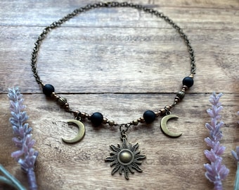 May include: A gold necklace with black beads and charms of a sun and two moons. The necklace is on a wooden surface with lavender flowers.