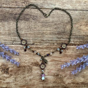 May include: A bronze necklace with a heart-shaped chain, featuring floral details and a mushroom pendant. The necklace is decorated with purple, brown, and clear beads. Lavender sprigs frame the necklace on a wooden surface.