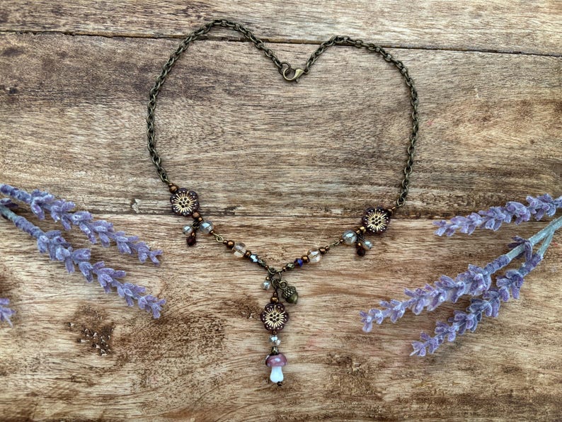 May include: A bronze necklace with a heart-shaped chain, featuring floral details and a mushroom pendant. The necklace is decorated with purple, brown, and clear beads. Lavender sprigs frame the necklace on a wooden surface.