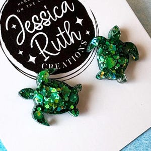 Può includere: Un paio di orecchini a lobo a forma di tartaruga marina verde con glitter incorporati, esposti su un cartoncino bianco con un logo circolare nero che riporta la scritta Jessica Ruth Creations Handmade on the Cape e piccole stelle.