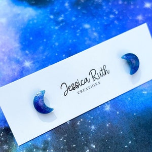 May include: A pair of crescent moon stud earrings with a blue and purple galaxy design. The earrings are on a white card with the text "Jessica Ruth Creations".