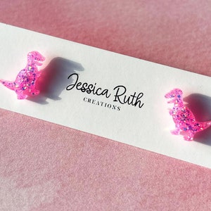 May include: A pair of pink glitter dinosaur stud earrings on a white card with the text "Jessica Ruth Creations".