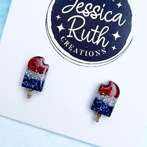 May include: A pair of popsicle-shaped stud earrings in red, white, and blue glitter resin. The earrings are on a white card with the text "Jessica Ruth Creations" and a navy blue circular logo. The background is light blue.