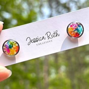 May include: Round stud earrings with colorful floral designs. The earrings feature a clear resin dome over a vibrant mix of pink, orange, yellow, green, and blue petals. The earrings are displayed on a white card with the text "Jessica Ruth Creations."